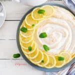 Pie de limón con decoración de rodajas de limón y hojas de menta, sobre plato azul y mesa de madera blanca con repasadores y taza de café